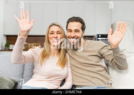 Écran vidéo avec un couple caucasien gai regardant la caméra et de l'ondulation. Webcam vue de la femme souriante et de son homme assis sur le canapé et salutation. Concept de réunion virtuelle Banque D'Images