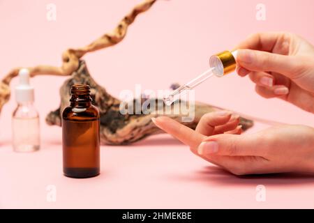 Une femme goutte du sérum facial d'une pipette sur son index. Gros plan des mains sur fond rose, décor naturel et cosmétique. Copier l'espace. T Banque D'Images