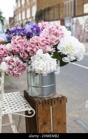 Un bouquet d'hortensias lilas, blancs et roses dans un seau en acier sur une boîte en bois est à vendre dans la rue à l'entrée d'un fleuriste Banque D'Images