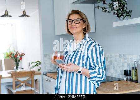Portrait d'une femme souriante d'âge moyen tenant son téléphone portable et buvant du thé tout en se tenant dans la cuisine moderne. Habitudes et rituels du matin. Jour p Banque D'Images