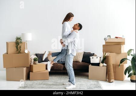 Un jeune couple aimant se réjouit dans leur nouvelle maison. Guy tient sa femme bien-aimée dans ses bras, dans le salon, ils ont déménagé dans un nouvel appartement, à côté de boîtes en carton avec des choses. Investissement immobilier Banque D'Images