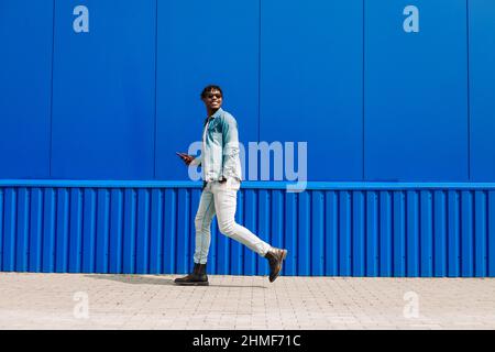 Pleine longueur d'un homme afro-américain élégant et gai en pleine croissance, utilise un smartphone et marche au-delà d'un mur bleu tout en allant au travail. L'homme tient Banque D'Images