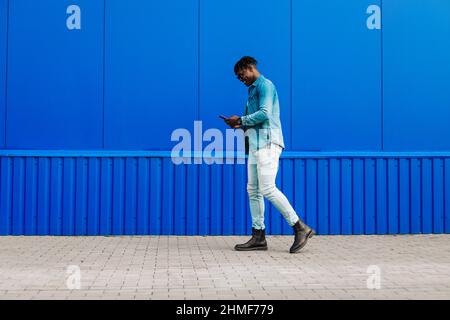 Pleine longueur d'un homme afro-américain élégant et gai en pleine croissance, utilise un smartphone et marche au-delà d'un mur bleu tout en allant au travail. L'homme tient Banque D'Images