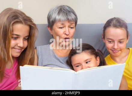 Grand-mère lit un livre avec des histoires à ses petites-filles assises ensemble sur le canapé Banque D'Images