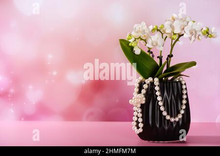 petite orchidée blanche dans un pot noir avec collier de perles, sur fond rose avec coeurs et sparkles Banque D'Images