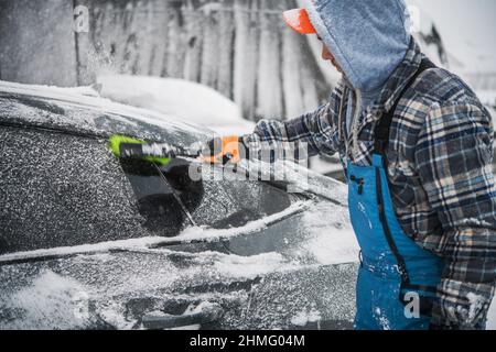 Les hommes caucasiens enlever la neige fraîche de sa voiture moderne à l'aide de l'outil de brosse douce. Banque D'Images