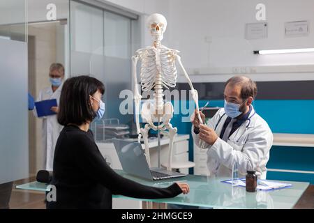 Médecin orthopédique expliquant la douleur des os sur le squelette humain au patient dans l'armoire médicale. Médecin pointant vers la moelle épinière sur le modèle d'ostéopathie pour montrer la structure anatomique et le diagnostic. Banque D'Images