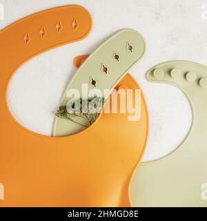 Composition de deux bavoirs en silicone orange et vert croisés avec fleur verte séchée sur fond blanc. Accessoires pour bébé, concept de première tétée. Mél Banque D'Images