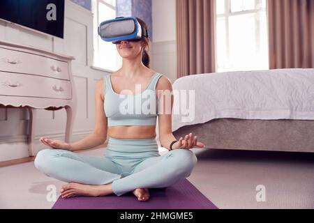 Femme portant un casque de réalité virtuelle assise sur un tapis de yoga dans la chambre à la maison et méditant Banque D'Images
