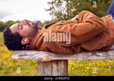 Homme allongé sur un banc à la campagne, se détendant et écoutant de la musique ou du podcast sur des écouteurs sans fil Banque D'Images