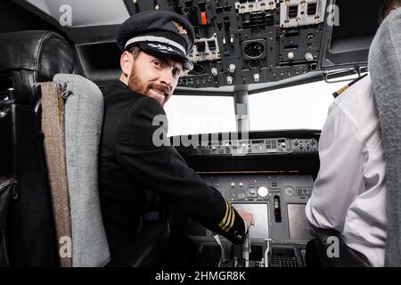 pilote barbu dans cap souriant et regardant la caméra près du copilote dans le simulateur d'avion Banque D'Images