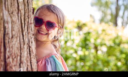 Jeune fille souriante portant des lunettes de soleil jouant Masquer et chercher derrière l'arbre dans le jardin Banque D'Images