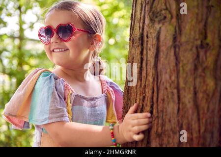 Jeune fille souriante portant des lunettes de soleil jouant Masquer et chercher derrière l'arbre dans le jardin Banque D'Images