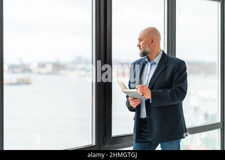 Homme d'affaires sérieux et mature en costume près de la fenêtre panoramique avec ordinateur portable Banque D'Images