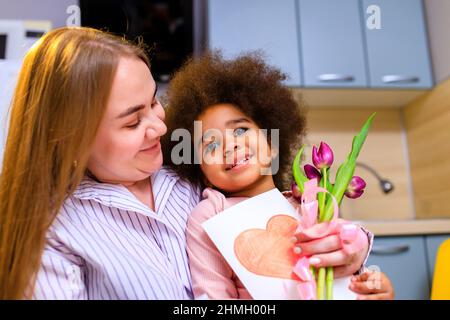 mère polyethnique et sa fille avec une coiffure afro assise dans la cuisine avec un bouquet de tulipes et une carte d'extension Banque D'Images