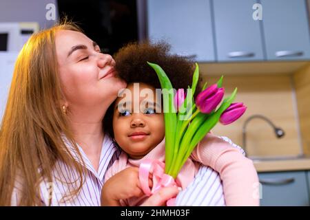 mère polyethnique et sa fille avec une coiffure afro assise dans la cuisine avec un bouquet de tulipes et une carte d'extension Banque D'Images