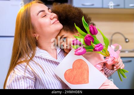 mère polyethnique et sa fille avec une coiffure afro assise dans la cuisine avec un bouquet de tulipes et une carte d'extension Banque D'Images