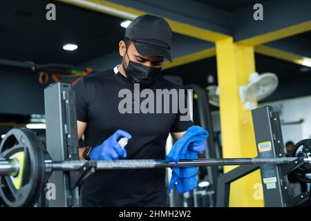 Jeune homme avec un masque médical et des gants de main aseptisant ou essuyant la sueur des équipements de gym en raison du coronavirus - concept de sécurité d'hygiène Covid-19 Banque D'Images