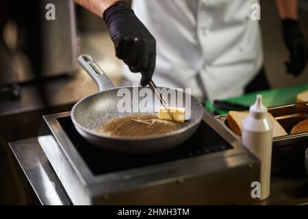 Vue en crêpe des mains du chef en gants noirs faisant fondre le beurre dans la poêle. Homme en uniforme de chef à l'aide d'une pince à épiler pour cuisiner avec des ingrédients et de l'équipement en arrière-plan. Concept de cuisine. Banque D'Images