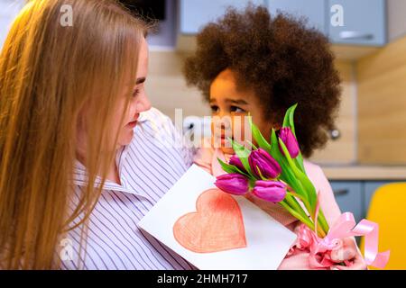 mère polyethnique et sa fille avec une coiffure afro assise dans la cuisine avec un bouquet de tulipes et une carte d'extension Banque D'Images