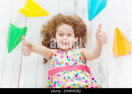 Rire fille jouer avec l'avion en papier et montrer les pouces vers le haut. Bonne enfance, voyage, vacances, concept d'éducation. Vue de dessus haute. Banque D'Images