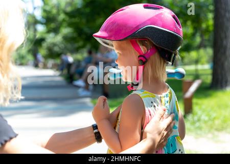 Gros plan maman parent mettre la main et attacher un casque de sécurité sur la mignonne blonde petite fille caucasienne pour faire du vélo ou scooter ville parc de rue à l'extérieur Banque D'Images