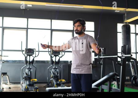 jeune constructeur indien de corps réchauffant le corps en sautant la corde avant de commencer l'entraînement à la salle de gym - concept de perte de graisse, soins du corps et bien-être. Banque D'Images