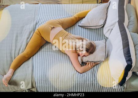 Femme aux cheveux longs blonds en haut jaune et leggings s'étire et se fend allongé sur le lit recouvert d'une couverture et d'un oreiller embrassant Banque D'Images