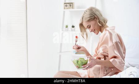 femme enceinte gaie tenant un bol avec salade et regardant le ventre dans la chambre Banque D'Images