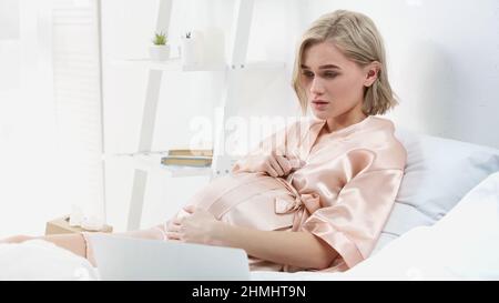 une femme enceinte blonde triste regardant un film sur un ordinateur portable dans la chambre Banque D'Images
