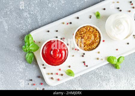 Ensemble de sauces populaires : ketchup, moutarde de dijon et mayonnaise dans de petits bols blancs sur un plateau en céramique vue du dessus Banque D'Images