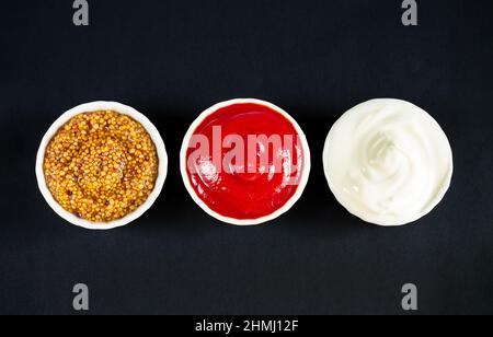 Ensemble de sauces populaires : ketchup, moutarde de dijon et mayonnaise dans de petits bols blancs sur fond noir. Vue de dessus Banque D'Images