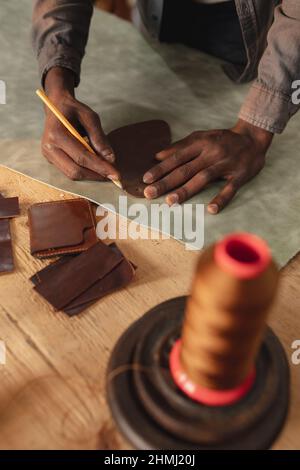 Section médiane de jeunes artisans afro-américains décrivant sur le cuir avec un crayon tout en faisant un portefeuille Banque D'Images