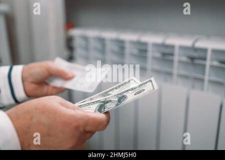 Des billets de dollars dans les mains des femmes près de la batterie de radiateur de chauffage à la maison.Frais de chauffage coûteux.Paiement des factures. Banque D'Images