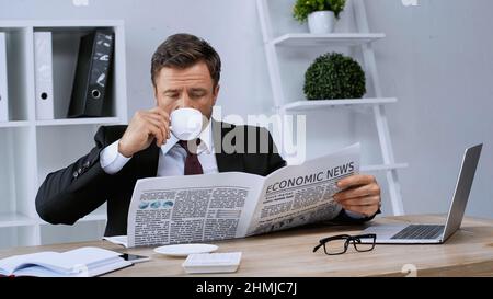 homme d'affaires qui boit du café et lit le journal près d'un ordinateur portable dans le bureau Banque D'Images