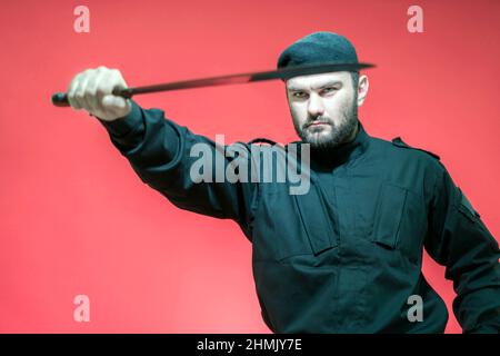 Défenseur des forces de l'ordre avec un regard époussin. Un gardien de sécurité mâle en uniforme noir et une casquette tient un long couteau dans sa main. Beau homme brutal. Mi Banque D'Images