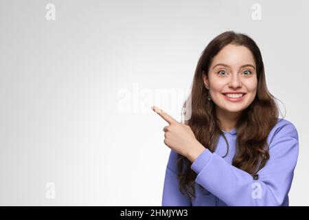 Une jeune femme aux cheveux longs et aux yeux bleus pointe son doigt vers la droite. La fille porte un pull à capuche violet surdimensionné Banque D'Images