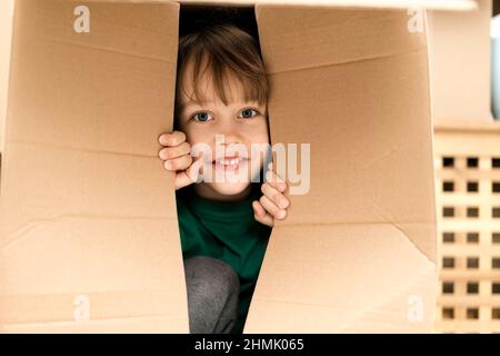 Garçon se cachant dans une énorme boîte en carton. Il joue et joue à travers un trou dans la boîte. Un enfant est heureux de s'installer dans une nouvelle maison. Banque D'Images