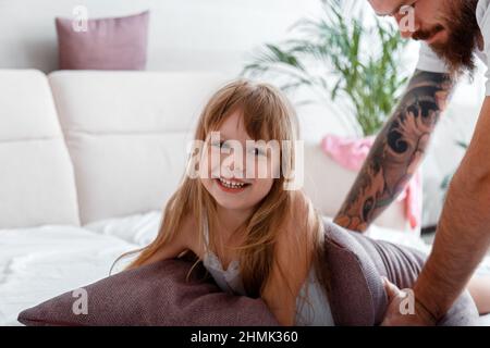 Portrait de l'enfant fille rires jouer avec papa. Père avec fille passant du temps ensemble jouer dans l'oreiller lutte dans le pyjama dans le lit. Chauve barbu Banque D'Images