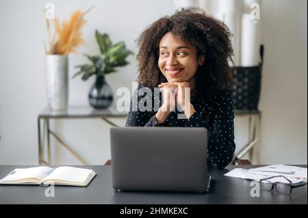 Jeune femme, employée de bureau, gestionnaire ou financier, à la peau sombre et satisfaite, assise à un bureau dans un bureau moderne, se repose sur le travail, prend une pause, regarde loin, sourit, rêver de repos Banque D'Images