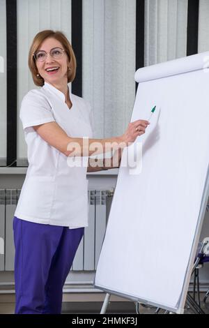 Préparation du projet. Femme souriante aux cheveux blonds en chemise médicale blanche qui écrit sur le paper-board Banque D'Images