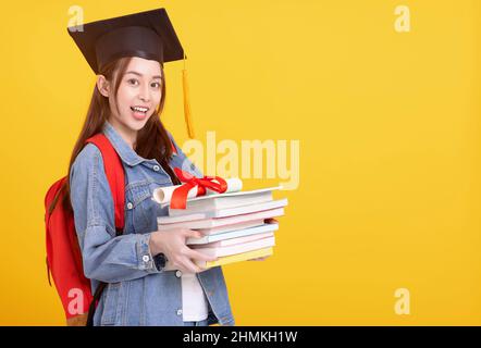 Bonne fille asiatique étudiant dans le chapeau de graduation souriant et tenant des livres Banque D'Images