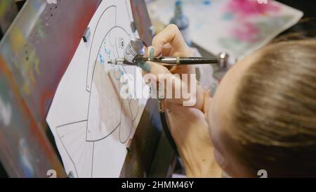 Une artiste apprend à peindre avec un aérographe avec du colorant acrylique, du papier et du chevalet. Intérieur. Concept art moderne, aérographe, aérographie, dessin pulvérisation image créer pulvérisateur dessin graffiti aérosol spray Banque D'Images