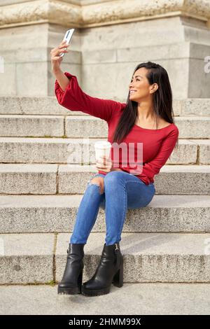 Jeune fille Latina prenant un selfie avec un téléphone cellulaire, se reposant sur les escaliers le jour du printemps. Latina en chandail rouge et jeans de détente dans la ville. Concept de style de vie. Banque D'Images