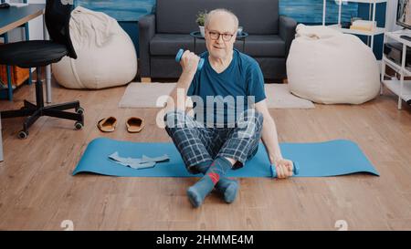 Homme aîné assis sur un tapis de yoga et des poids de levage pour former des muscles à la maison. Retraité utilisant des haltères pour faire de l'exercice physique et étirer les bras, de l'entraînement et de faire de l'activité d'entraînement. Banque D'Images