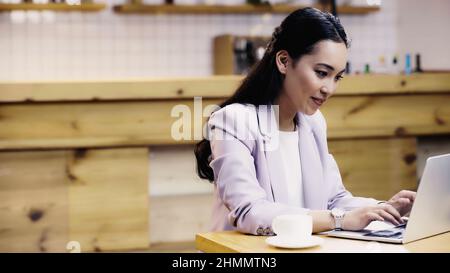 jolie femme d'affaires asiatique en costume de taper sur un ordinateur portable tout en travaillant à distance dans un café Banque D'Images