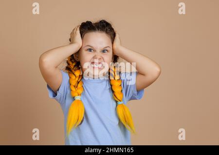 Colère offensé contrarié caucasien petite fille tenant la tête grinning dents regardant la caméra ayant kanekalon braides de couleur jaune dans le t-shirt bleu sur Banque D'Images