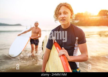 Jeune garçon blond adolescent avec un père portant des planches de surf alors qu'ils marchent sur la plage de sable de l'océan après avoir surfés sur fond de beau coucher de soleil. Ils le sont Banque D'Images