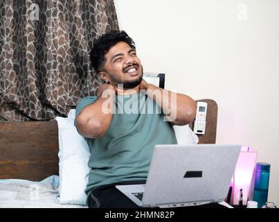 jeune homme souffrant de douleurs au cou et au dos tout en utilisant l'ordinateur portable sur le lit pendant le travail de la maison - concept de la posture de travail incorrecte, surtravaillé et Banque D'Images