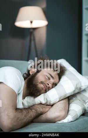 Beau jeune homme barbu dormant sur le canapé en embrassant un oreiller après de longues heures de travail tard le soir. Jeune homme malade dormant sur le canapé après avoir pris des médicaments. Concept de travail à distance. Banque D'Images